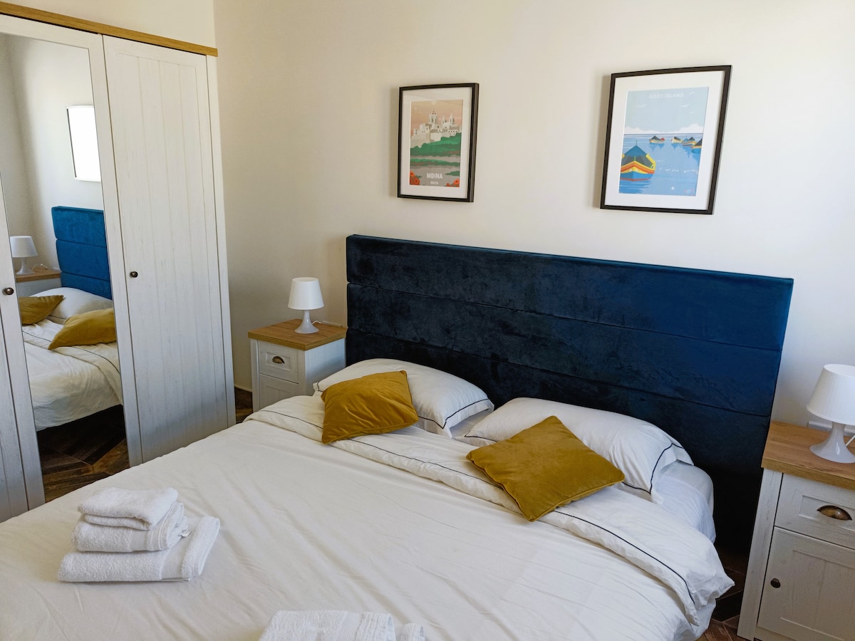 A cozy bedroom is highlighted by a spacious bed adorned with soft pillows and golden accent cushions. Complimentary white towels are neatly arranged on the bed. The room features a wardrobe and bedside tables, each with a lamp, and two framed artworks are displayed on the wall.