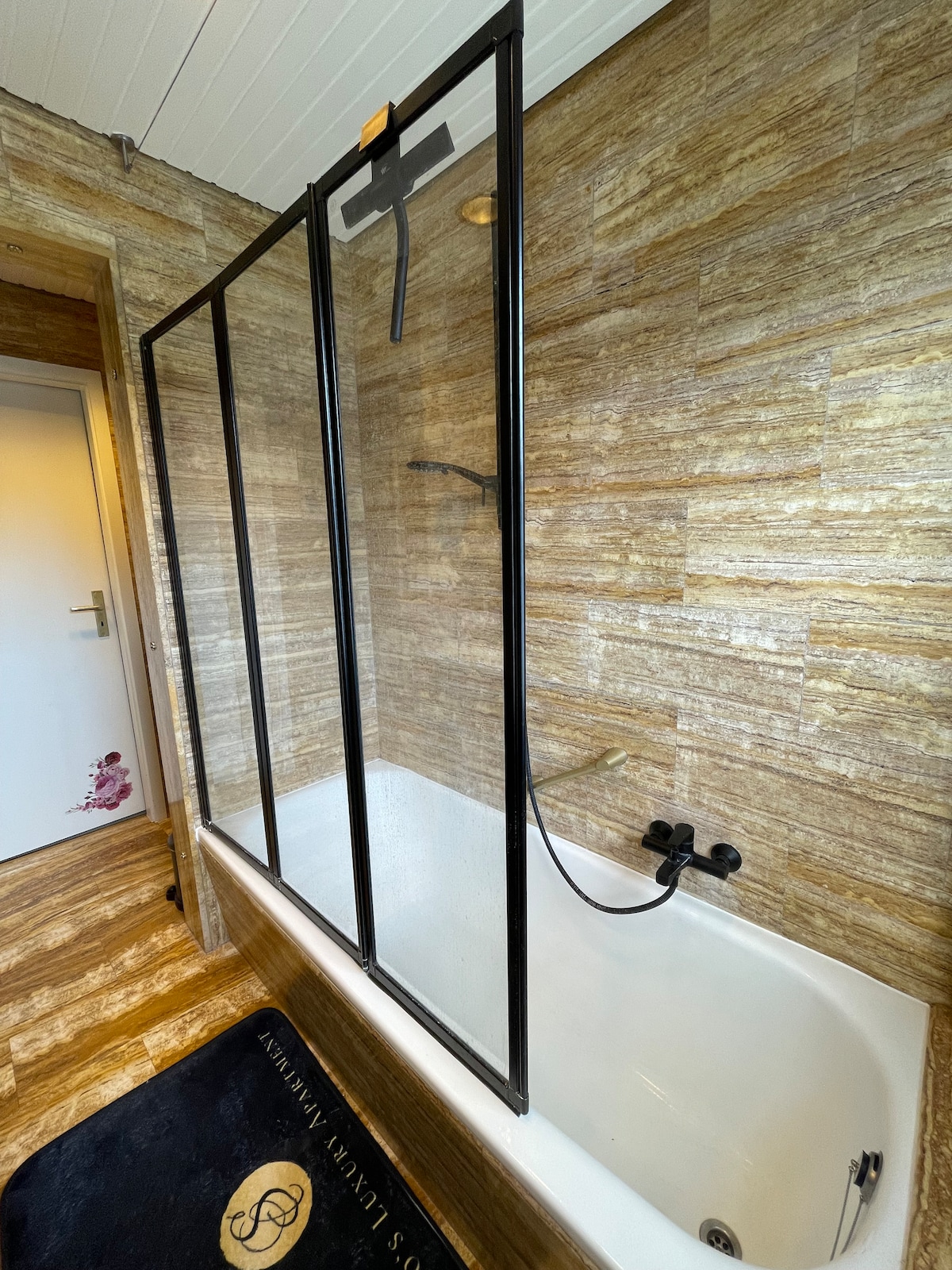 A modern bathroom features a freestanding bathtub enclosed by a glass shower screen. The walls are adorned with textured stone tiles, and a sleek black shower fixture is visible. A dark rug lies on the floor, adding a touch of contrast to the overall design.