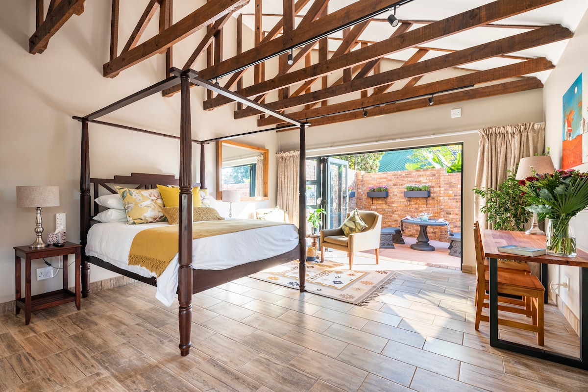 A spacious bedroom features a four-poster bed adorned with soft linens and pillows. Wooden beams span the ceiling, enhancing the open feel. A sliding glass door leads to a private patio with outdoor seating. A small dining table and decorative flowers are present.