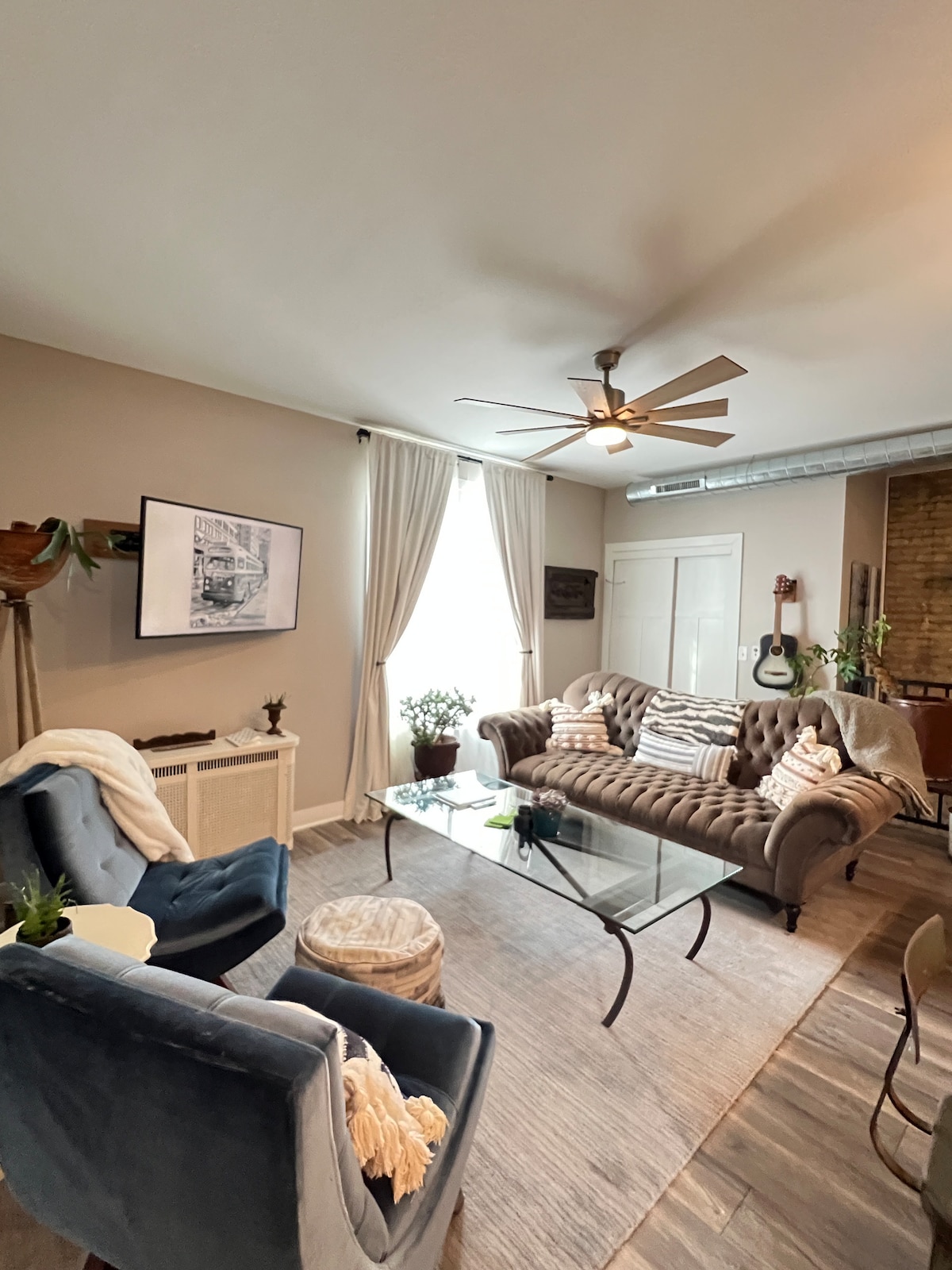 A cozy living area features a tufted sofa and two upholstered chairs, complemented by a glass coffee table. Natural light enters through a large window adorned with light curtains. Various plants and decorative items add charm to the space, enhancing its inviting atmosphere.
