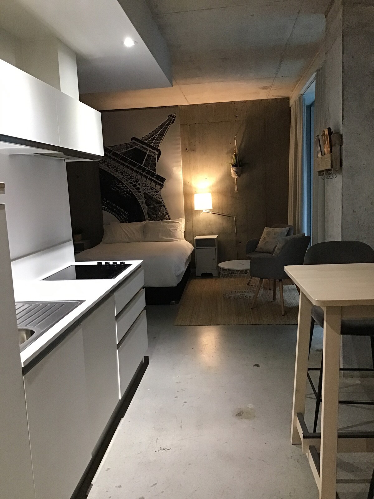 A compact living space is presented, featuring a well-equipped kitchen with white cabinetry and a sleek countertop. A cozy sleeping area is highlighted with a large mural of the Eiffel Tower. A comfortable armchair and a small table create a relaxing nook beside the bed.