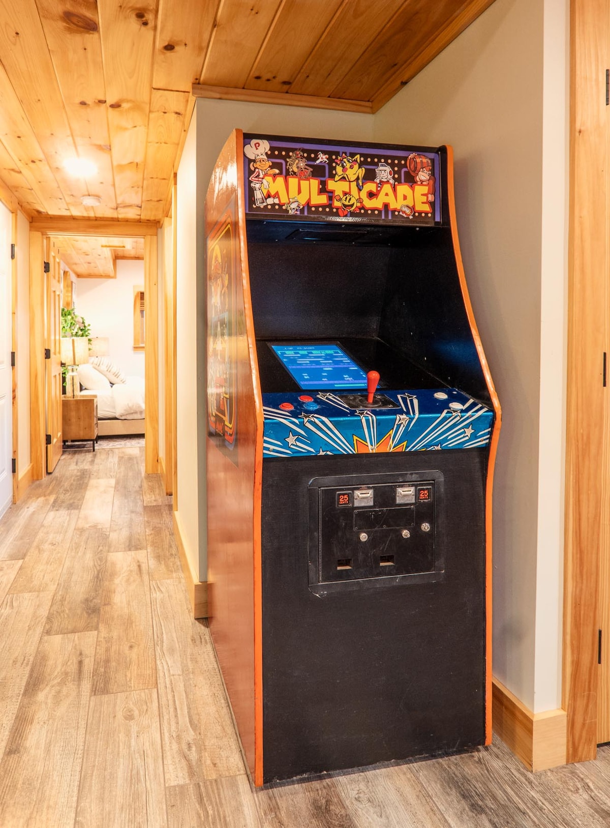 A vintage-style arcade game machine is positioned in the hallway, featuring colorful artwork and a bright screen. The hallway is accented with natural wood finishes and a warm ceiling, leading to additional areas within the cabin.