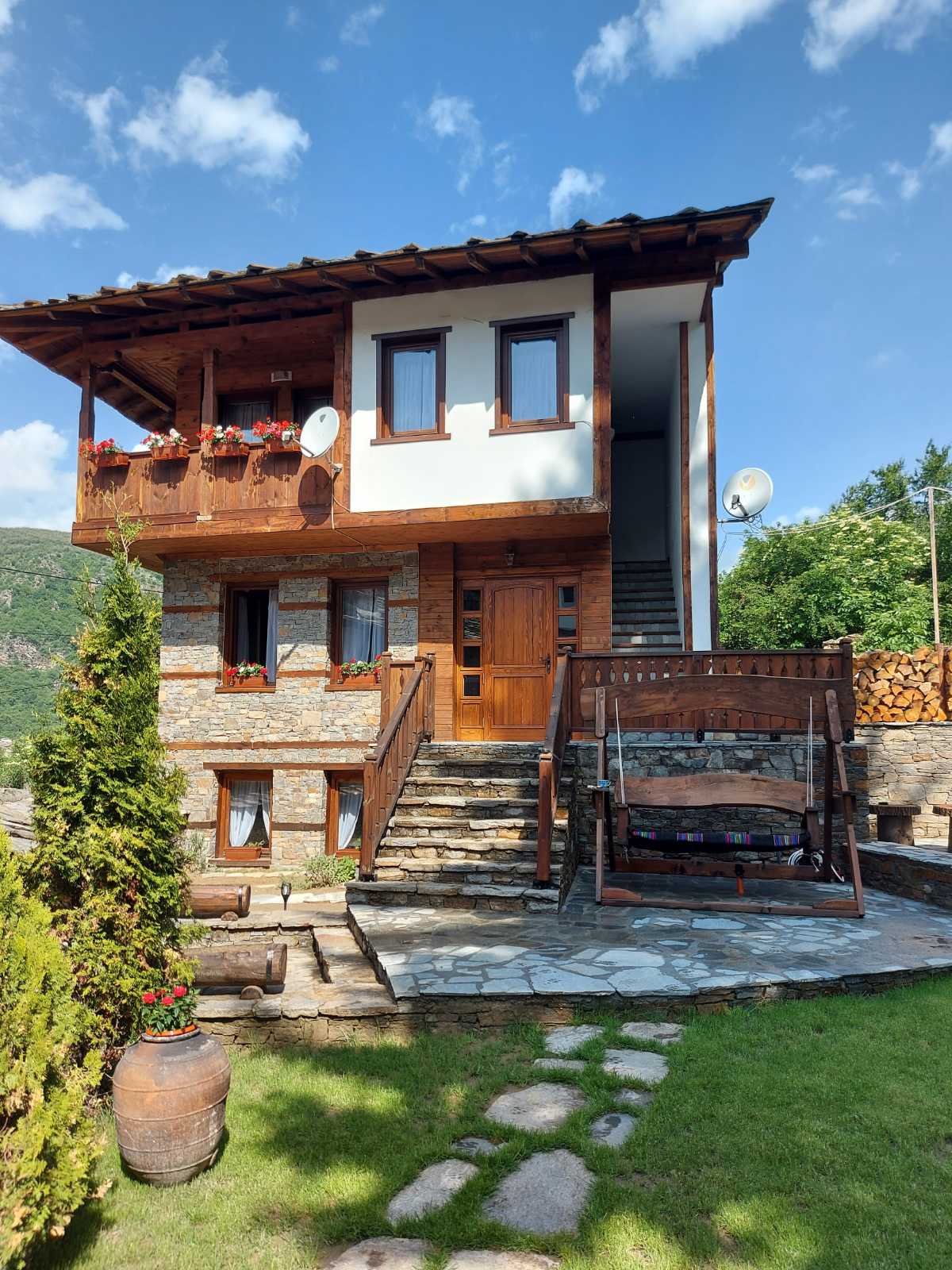 The two-story stone and wooden house is surrounded by a well-maintained garden. A stone pathway leads to the entrance, framed by blooming flowers. A wooden swing sits on the patio, with large windows allowing natural light to brighten the interior spaces.