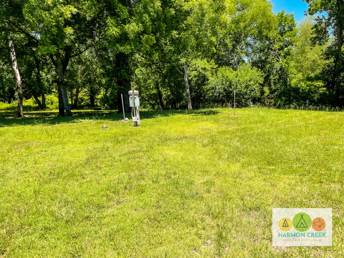 A spacious grassy area is surrounded by lush trees, providing shade and natural beauty. Utility hookups for RVs are visible on one side, offering easy access for connections. The site is level and clear, creating an inviting space for outdoor activities.