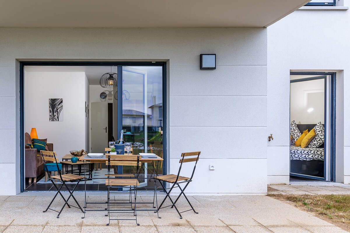 A private terrace offers a dining table with four wooden chairs, surrounded by a grassy area. Glass doors provide a view into an interior space featuring a cozy living area with soft seating and art. A well-lit ambiance is created by natural light.