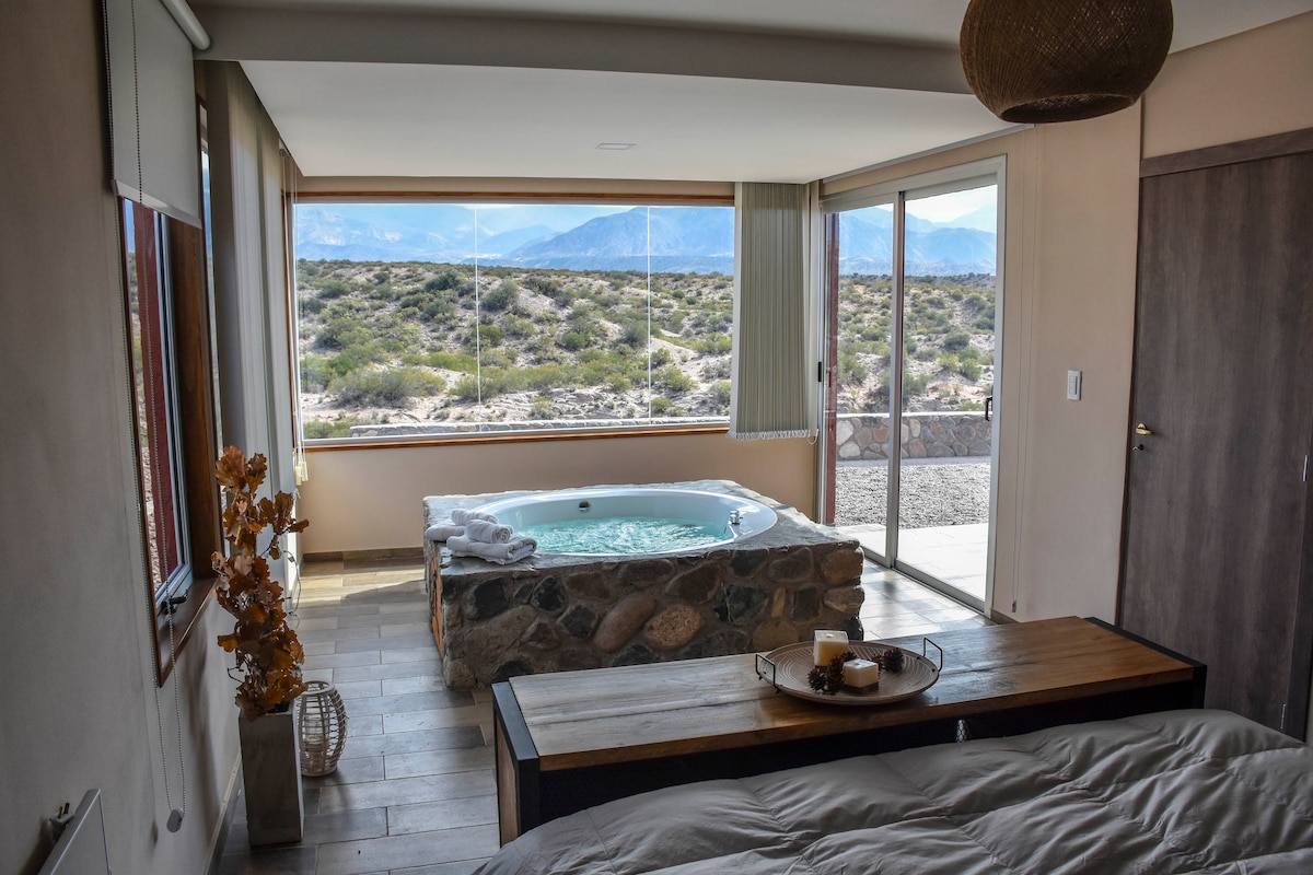 A spacious bedroom features a large window overlooking the scenic landscape, allowing natural light to fill the space. A cozy bed is positioned nearby, and a hot tub is situated on the stone platform, enhancing the serene atmosphere of the room.