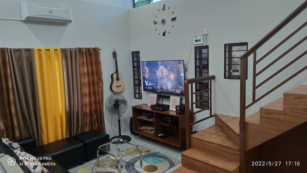 A cozy living area is featured, showcasing a dark sectional sofa and a round, patterned rug. A wall-mounted television is visible, accompanied by a wooden entertainment unit. Natural light enters through large windows, with colorful curtains adding warmth to the space. A staircase leads to the upper level.