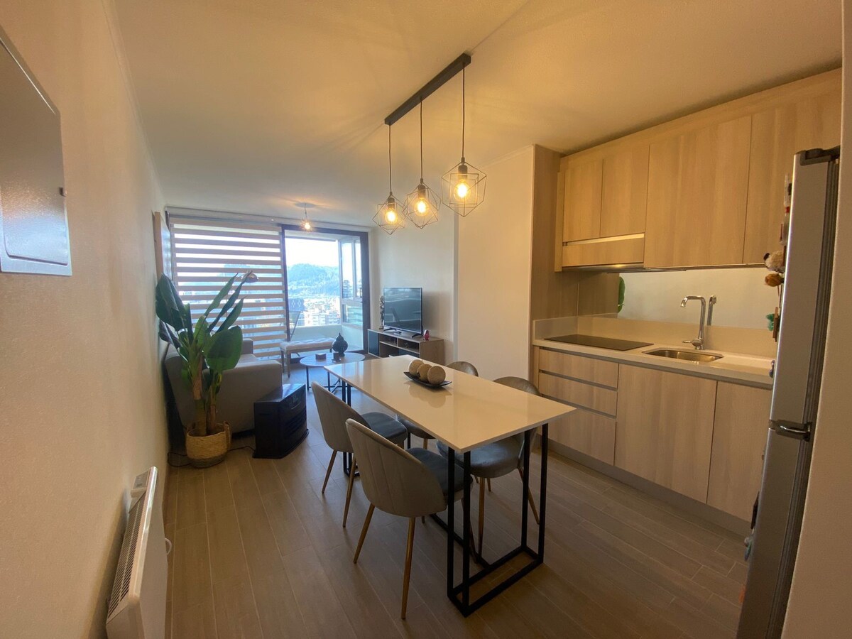 A dining area is presented with a modern table and four chairs, complemented by a sleek kitchen with wooden cabinetry. A large window offers views of the outside, while a small indoor plant is positioned near the corner, adding a touch of greenery.