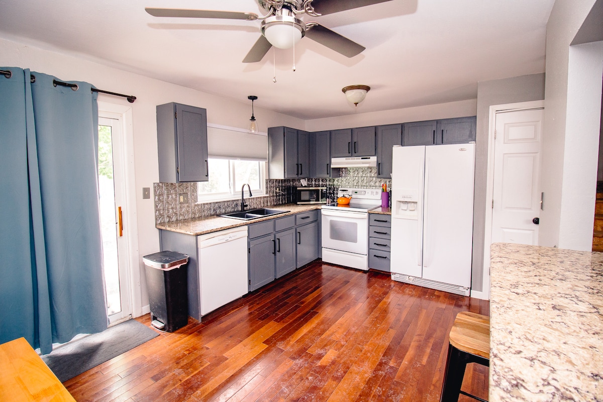 The kitchen features light gray cabinetry paired with a granite countertop. A ceiling fan provides airflow, while a white refrigerator and stove are easily accessible. Natural light enters through a window, illuminating the wooden floor and enhancing the spacious layout.