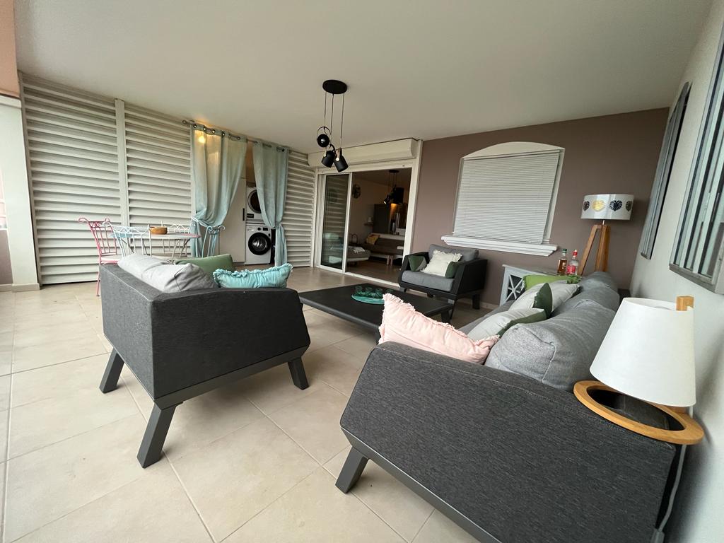 A spacious living area features comfortable seating with gray sofas and decorative pillows. A small dining table is visible in the background, while light and airy curtains enhance the natural light. The area has a modern aesthetic, with a neutral color palette and minimal decor.