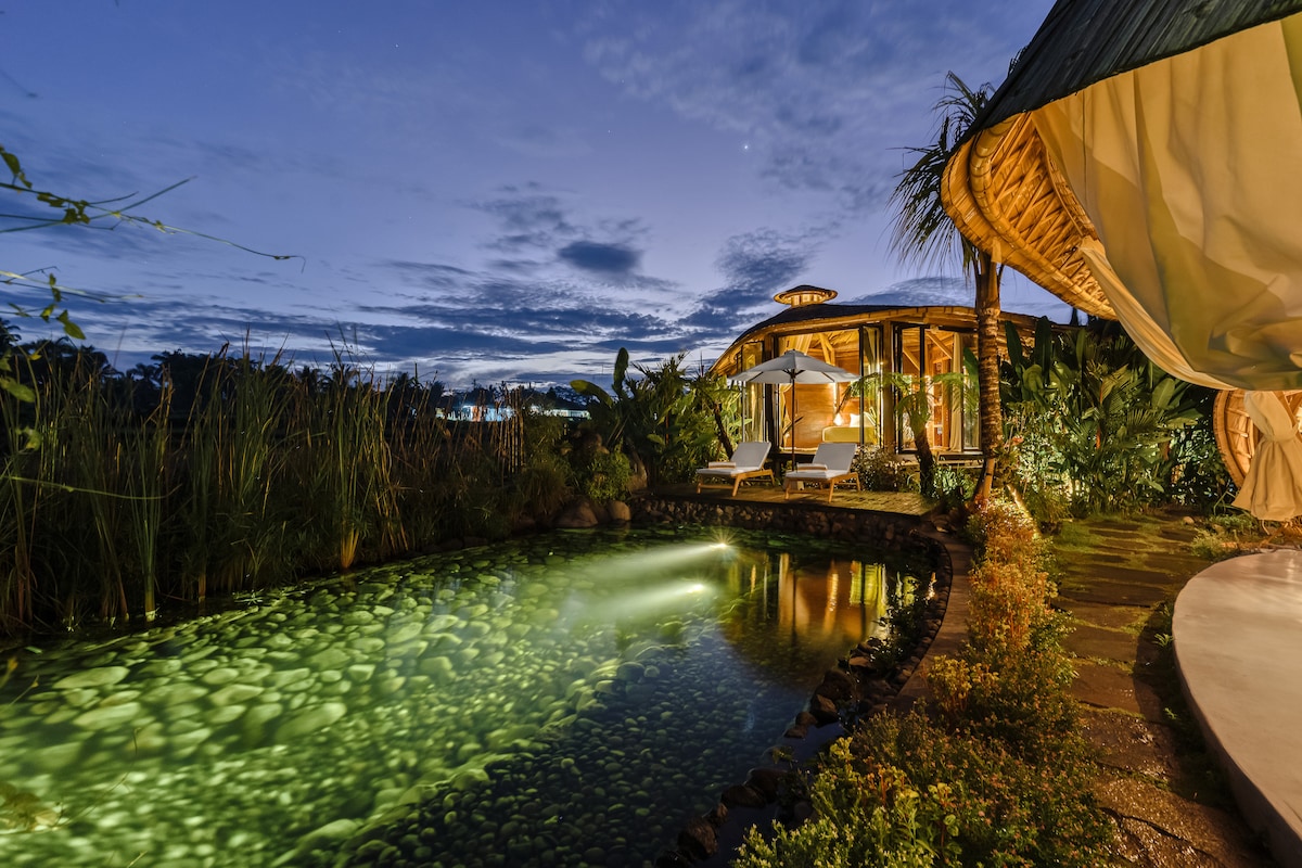 A serene natural pool glows softly in the evening light, surrounded by lush greenery. Comfortable lounge chairs are positioned beside the water, offering a tranquil space for relaxation. The villa's design harmonizes with the landscape, contributing to an inviting atmosphere for guests.