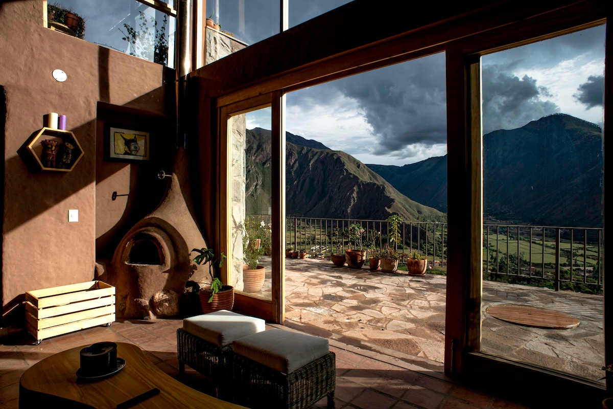 A spacious living area features large glass doors that open onto a stone terrace, offering panoramic views of the mountains and valley. A rustic fireplace with a clay exterior stands prominently, while potted plants add a touch of greenery to the interior.