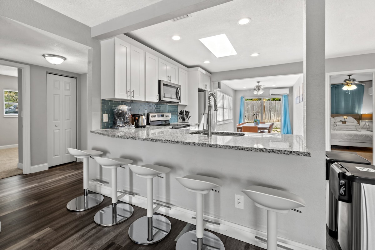 The modern kitchen features a spacious island with sleek bar stools, complemented by stainless steel appliances and white cabinetry. Natural light is provided through a skylight, illuminating the open floor plan that connects to a dining area, creating an inviting space for meals and gatherings.