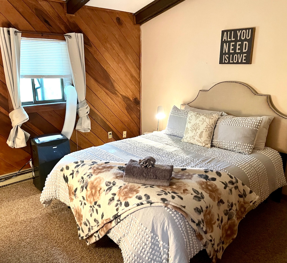 A comfortable bedroom features a spacious bed adorned with a floral blanket and a hand towel. The walls are accented with natural wood paneling. Light enters through a nearby window, framed by soft curtains, while a decorative wall sign adds a personal touch.