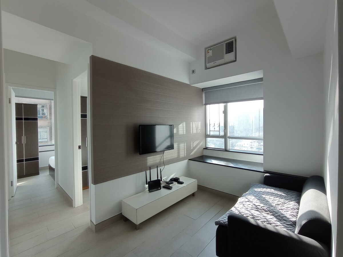 A bright living area features a wall-mounted television above a modern white media console. A black sofa offers seating, while large windows allow natural light to fill the space, revealing views of the city outside. The room exhibits a minimalist design aesthetic.