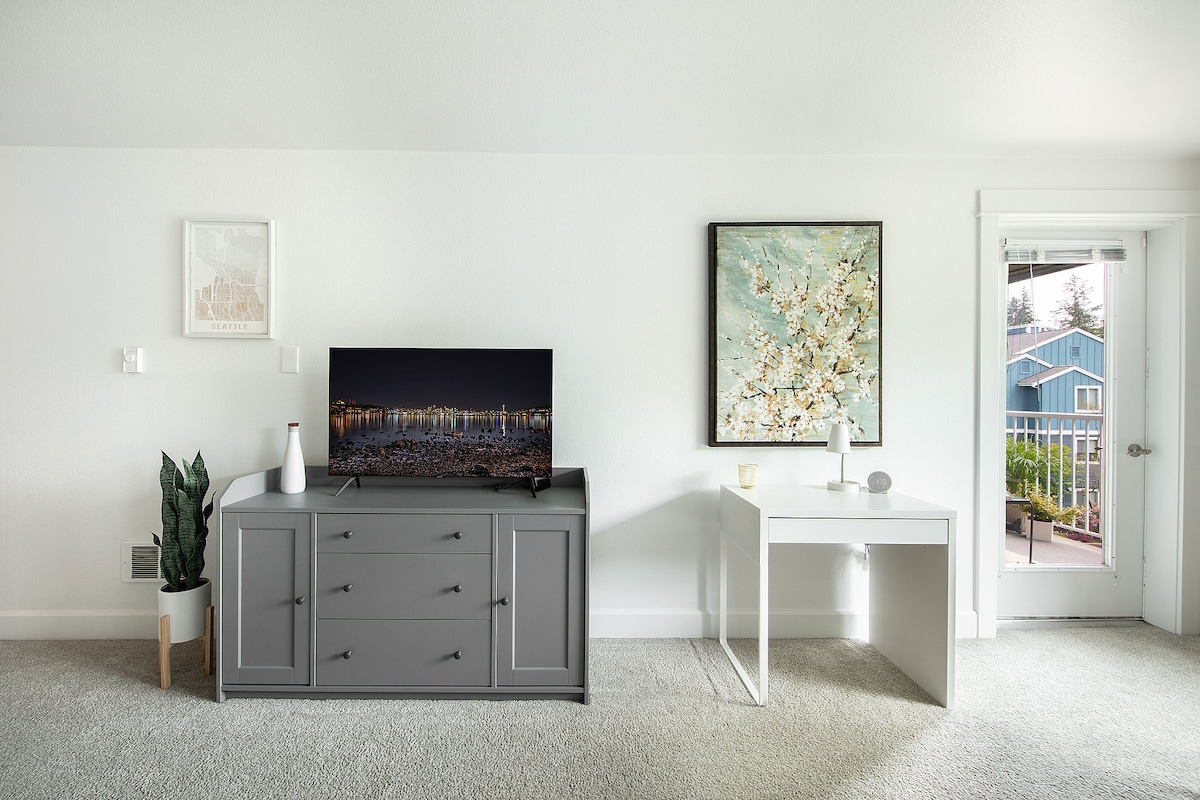 A bright and spacious living area features a television on a gray media console, complemented by a decorative plant nearby. A white desk offers a functional workspace, while a door leads to an outdoor deck. Art pieces adorn the walls, enhancing the tranquil environment.