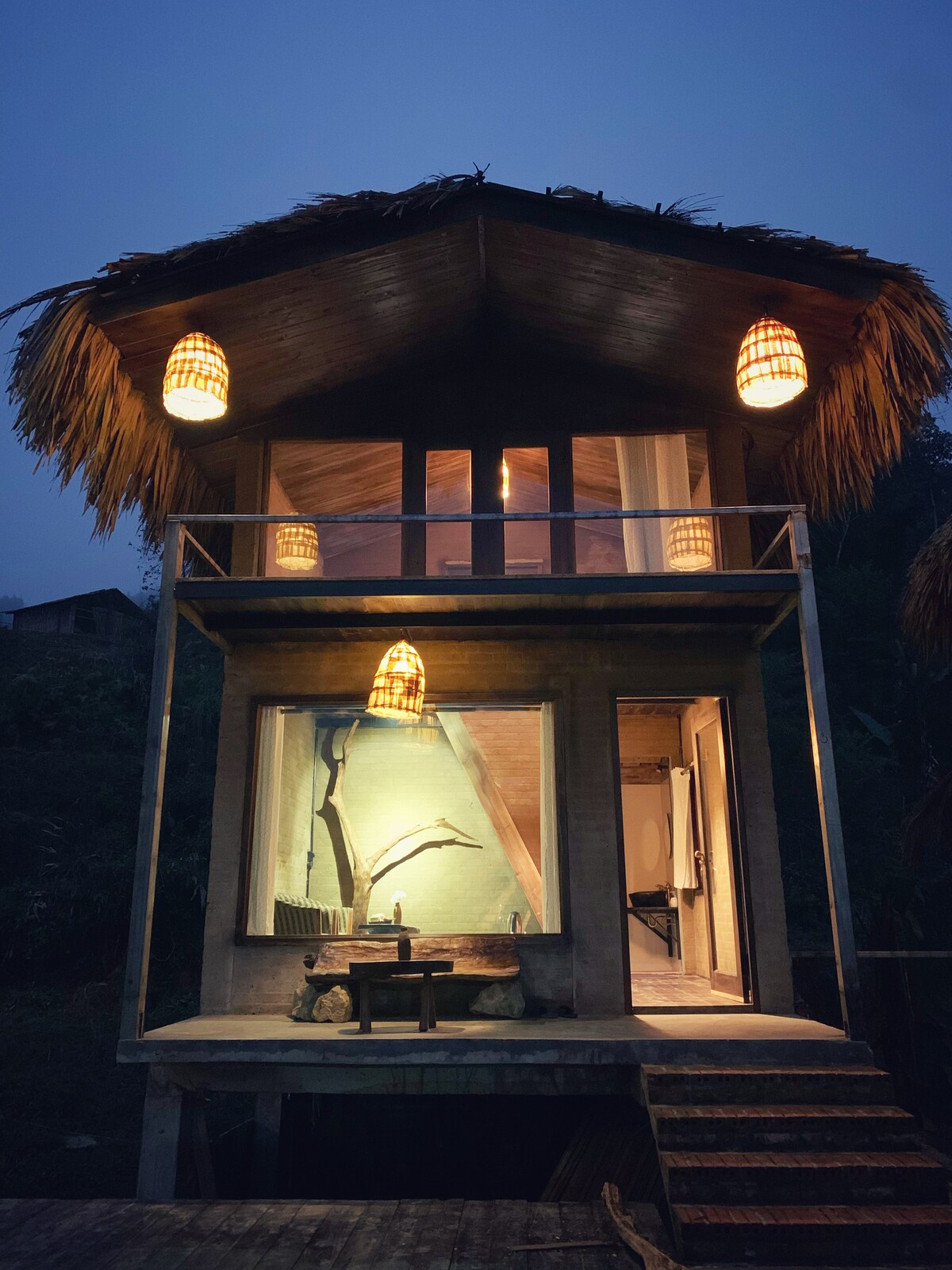 The illuminated chalet bungalow is showcased at dusk, with soft lighting highlighting its wooden features. A cozy balcony overlooks the surroundings, while a warmed interior is visible through the large windows, presenting a rustic charm that blends with the natural environment.