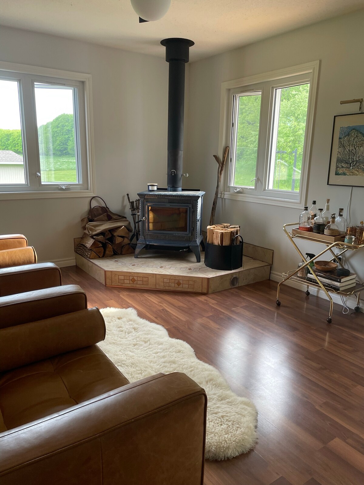 A cozy corner features a wood stove positioned on a stone platform with a stack of firewood nearby. Two brown leather chairs are placed on polished wooden flooring, while large windows allow natural light to fill the space, offering views of the surrounding greenery.