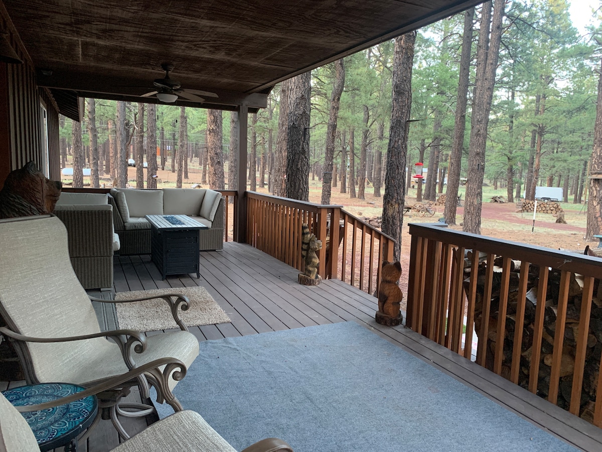 A front porch is displayed, featuring a blend of comfortable seating including two armchairs and a sectional sofa. A wooden railing enhances the view of surrounding trees, while a small coffee table stands at the center. A rug and decorative totems add warmth to the space.