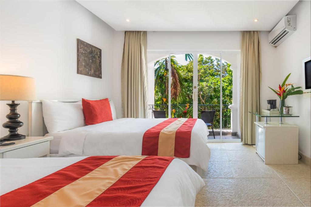 An elegantly decorated room features two queen-size beds dressed in crisp white linens with red and gold accents. A glass door opens to a furnished balcony surrounded by lush greenery. A nightstand with a lamp and a modern coffee station are visible.