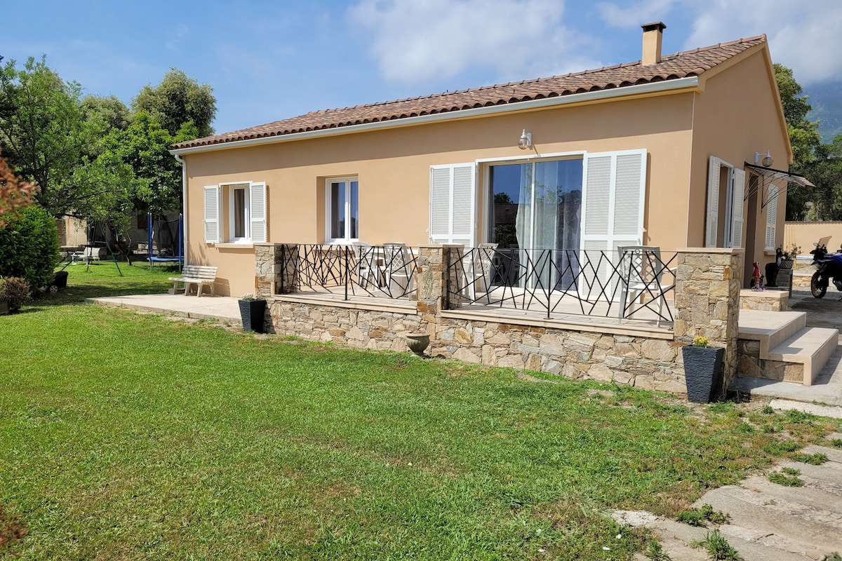 A single-story villa features a light-colored facade and extends to a spacious terrace. Large glass doors open to the outside. A well-maintained lawn surrounds the structure, with decorative planters placed near the entrance, creating an inviting outdoor environment.
