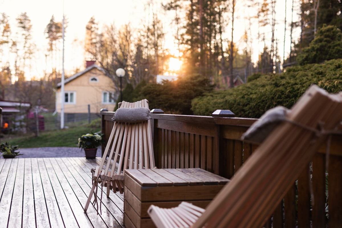 A wooden deck features two reclining chairs facing a serene garden. Lush greenery surrounds the space, and soft sunlight filters through the trees, creating a warm ambiance. Decorative plants are placed nearby, enhancing the inviting outdoor area.