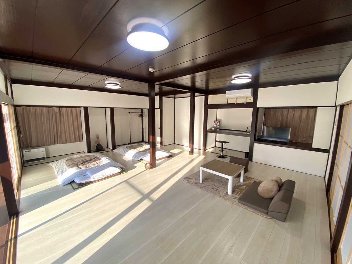 A spacious interior features wooden beams and ample natural light. Two comfortable futons are neatly arranged on the floor, complemented by a low table and a cushioned seating area. Large windows offer a view of the outdoors, enhancing the tranquil atmosphere.