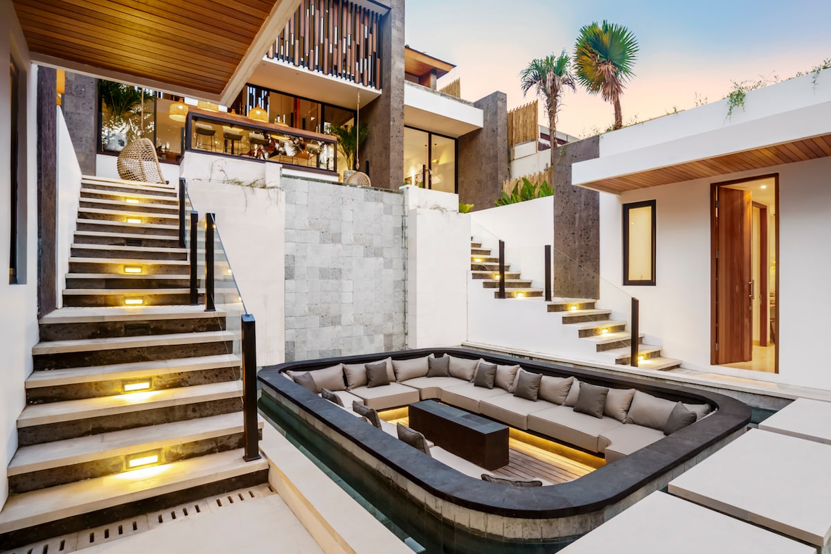 A contemporary outdoor space features a sunken lounge area surrounded by a sleek pool. Stairs lead up to the villa, illuminated by integrated lights. The design incorporates natural materials and greenery, creating a seamless connection between the architecture and the landscape.