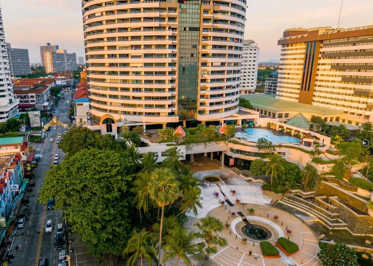 The exterior view showcases a modern high-rise building with a curved shape, surrounded by palm trees and lush greenery. A large swimming pool is visible, with lounge areas beneath umbrellas. The surrounding urban landscape features a mix of buildings and a road lined with trees.