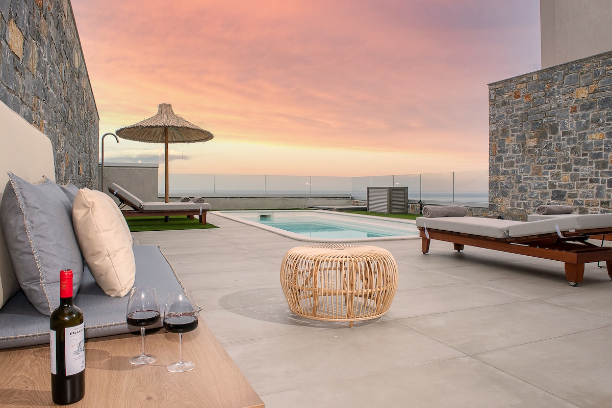An outdoor seating area features a comfortable lounge with soft cushions and a round wicker table. Two wine glasses and a bottle are placed on the table. The space is bordered by stone walls, leading to a private pool reflecting the colorful sunset sky.