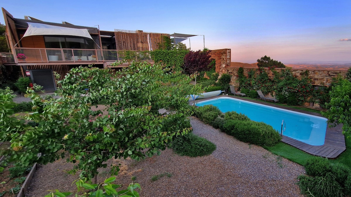 A tranquil outdoor area features a swimming pool surrounded by neatly landscaped gardens. The pool, framed by lush greenery, reflects the sky, while the main building, adorned with wooden elements, rises in the background, offering a serene ambiance in nature.