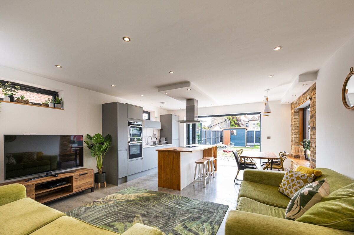 An open plan living area is showcased, featuring a modern kitchen with sleek cabinetry and a spacious island. Natural light floods in through bi-fold doors, which connect to a sunny outdoor space. Comfortable seating is arranged, creating an inviting area for relaxation.