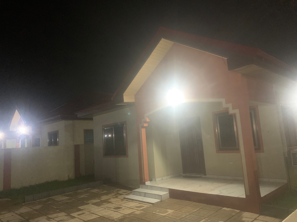 A single-story building is showcased at night, featuring illuminated exterior lights that enhance its architecture. The entrance is marked by a spacious porch with steps leading up to a wooden door. Smooth tiles create a welcoming path in front of the home.