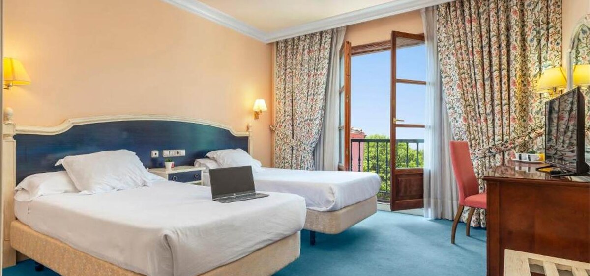 A modern hotel room features two expansive beds positioned opposite a wooden desk with a laptop. Large floral-patterned curtains frame the window, which opens to scenic mountain views. A flat-screen TV is mounted on a wooden dresser, complemented by a single chair.