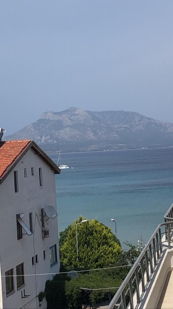 A scenic view showcases a mountain rising above the serene waters, with varying shades of blue visible in the sea. Nearby buildings are seen in the foreground, with a mix of greenery adding to the landscape. The atmosphere appears tranquil and inviting.