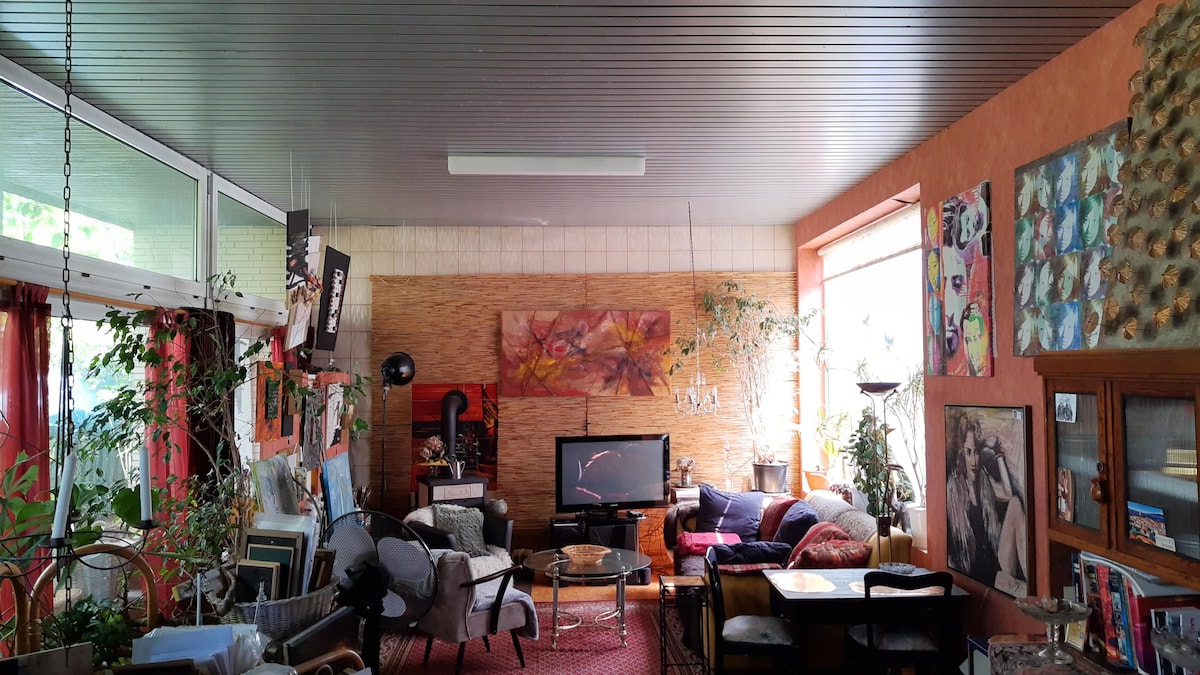 A spacious living area is visible, featuring a mix of comfortable seating options, including a large sofa and several armchairs. A television is positioned in front of a fireplace. Various artworks adorn the walls, and potted plants add a touch of greenery throughout the room.