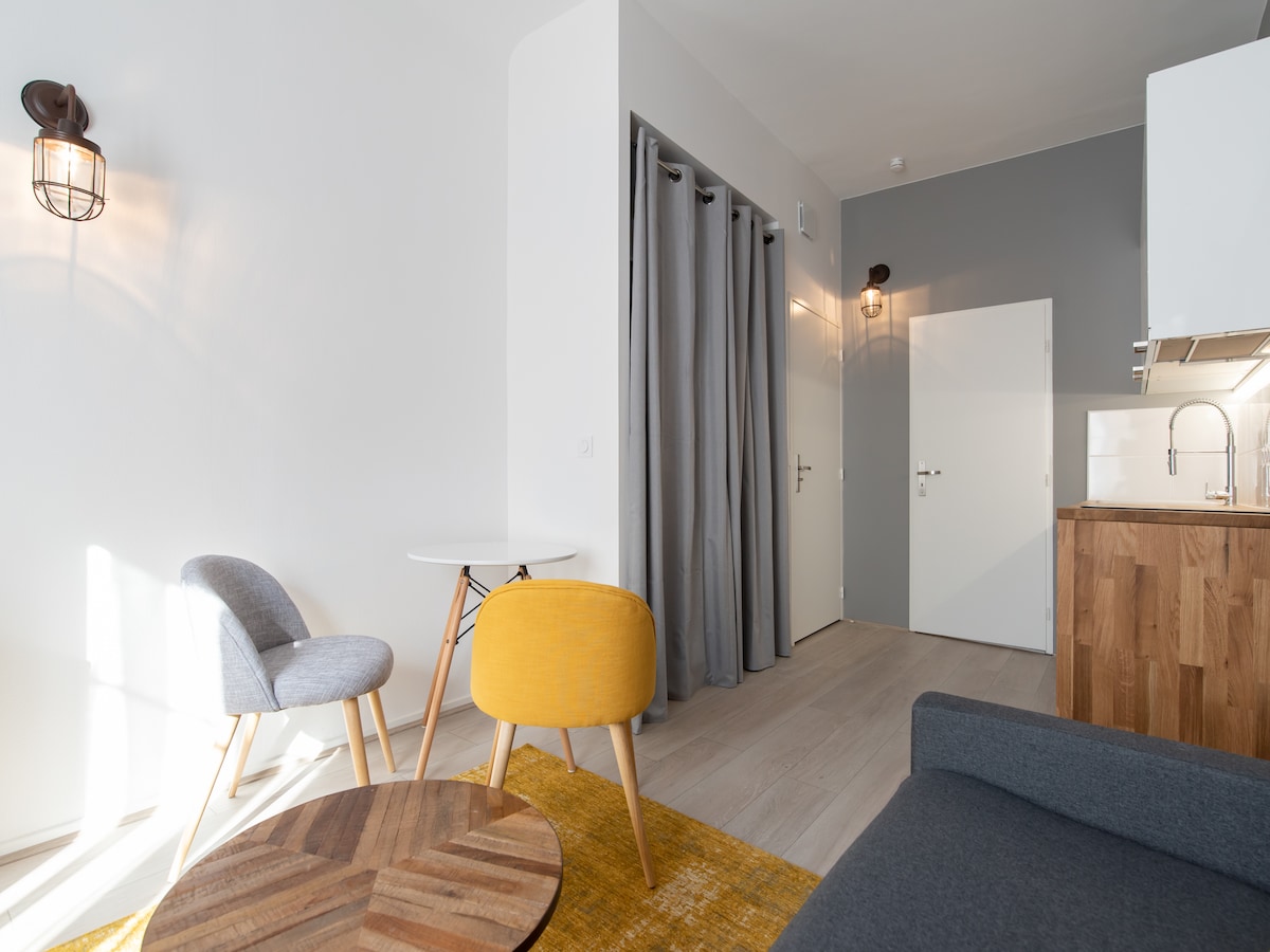 A modern living area features a grey sofa, a round wooden coffee table, and two accent chairs in grey and yellow. Light enters through a window, casting gentle shadows on the wall. The space is paired with a sleek kitchen, partially visible.
