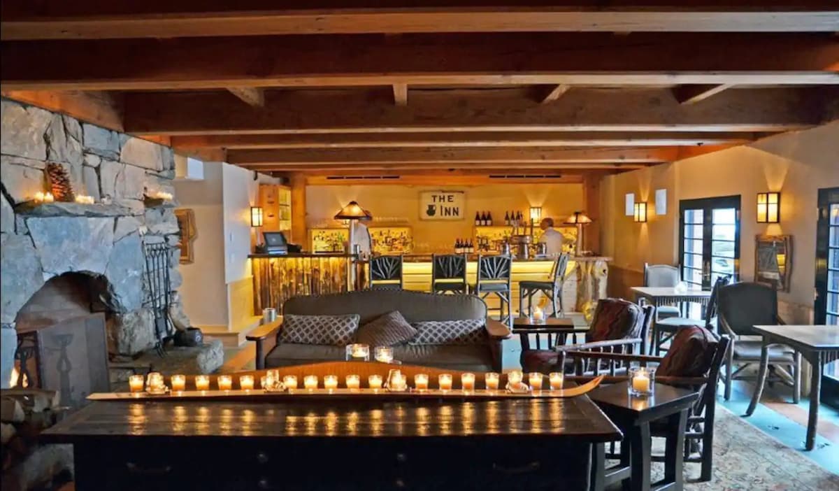 A warm and inviting lounge area features exposed wooden beams and a stone fireplace. Soft seating options are arranged in front of a low table, which is adorned with flickering candles. The bar area is visible in the background, showcasing a welcoming atmosphere.