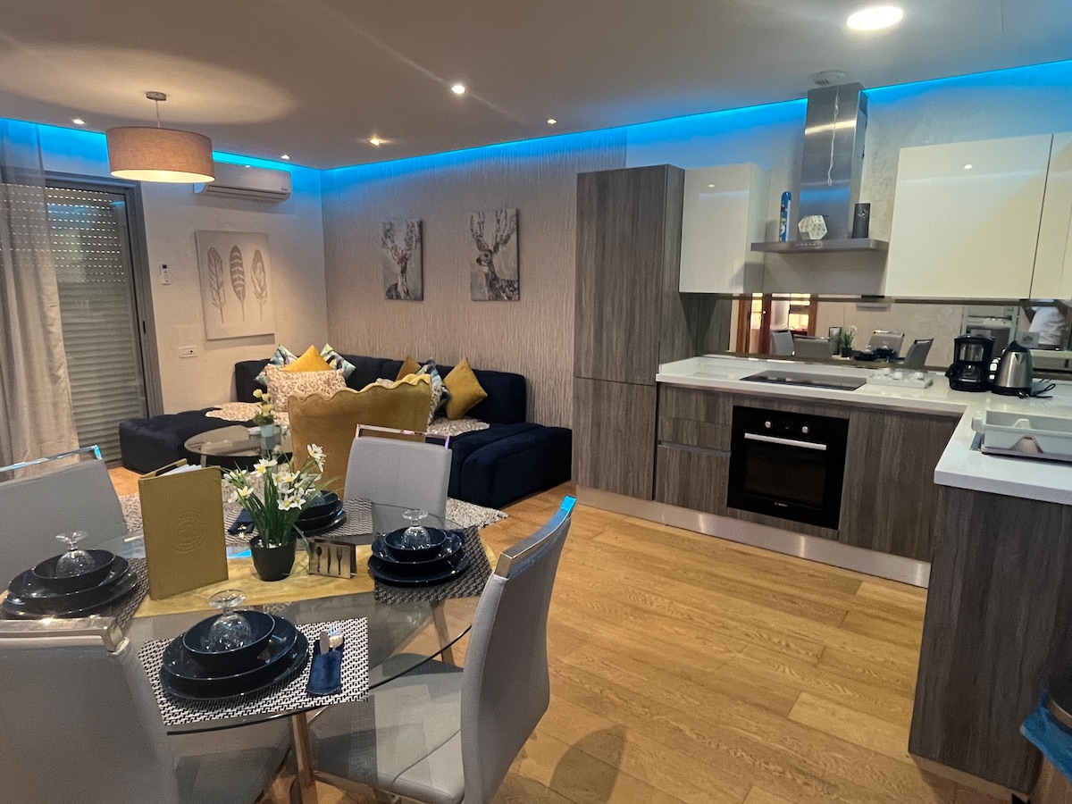An open-plan living area features a modern kitchen with sleek cabinetry and stainless steel appliances. A dining table with grey chairs is set elegantly, while a cozy lounge space displays navy and yellow accents. Soft lighting creates a warm ambiance, complementing the wooden flooring.