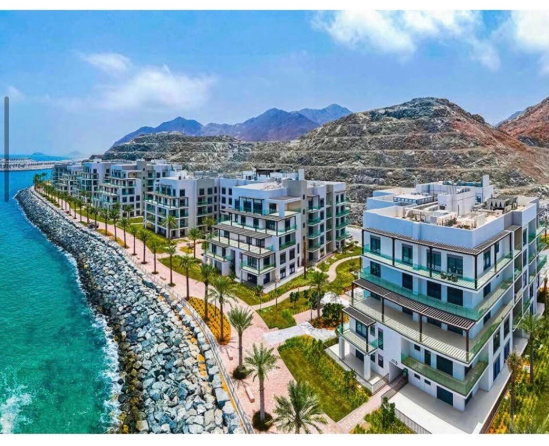 A serene coastal view features modern buildings lining a pathway along the water's edge, surrounded by lush greenery and palm trees. The landscape includes rocky shorelines and distant mountains under a clear blue sky, creating a peaceful atmosphere.