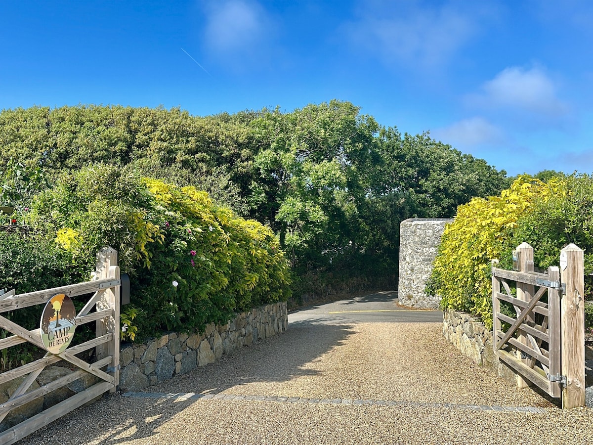 The entrance to Camp de Rêves Glampsite is framed by dense greenery and vibrant foliage. A wooden gate, adorned with a welcoming sign, opens onto a gravel driveway lined with lush plants, leading towards the serene and spacious glampsite.
