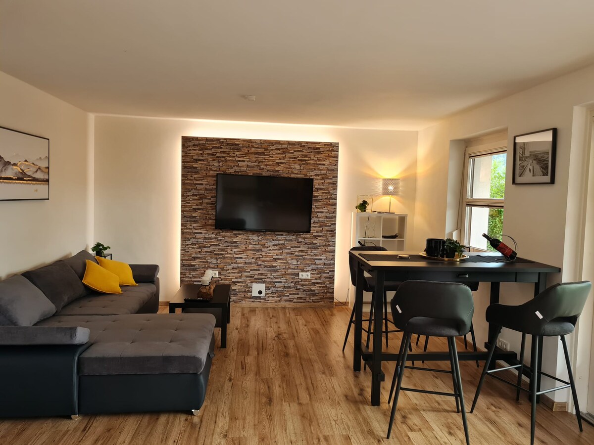 A modern living area features a spacious sectional sofa complemented by a yellow throw pillow. A flat-screen television is mounted on a textured stone wall, while a dining space with high-backed chairs and a small table invites casual meals. Natural light floods the room through large windows.