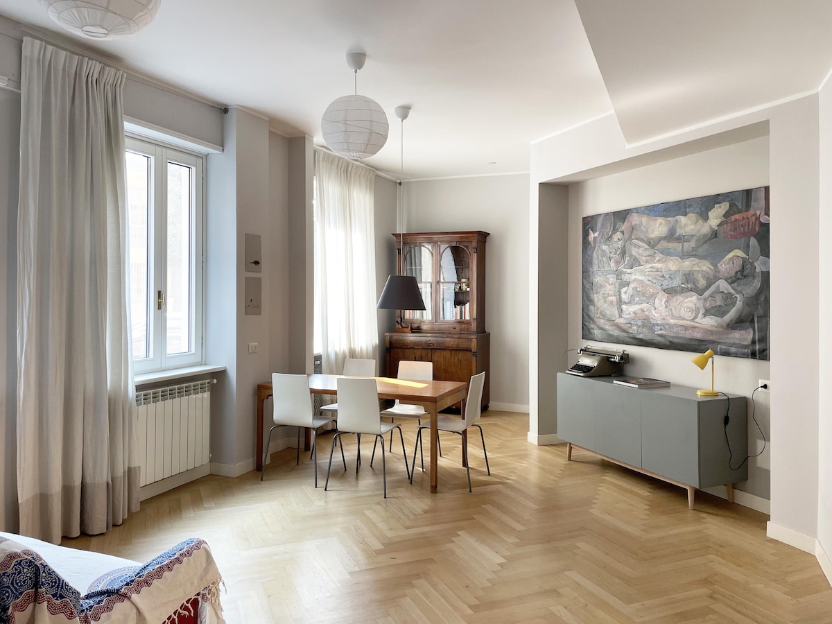 A bright living area features light-colored walls and warm wooden flooring. A dining table with four chairs sits in the center, accompanied by a vintage cabinet and a modern sideboard. Natural light enters through large windows, enhancing the spacious feel of the room.