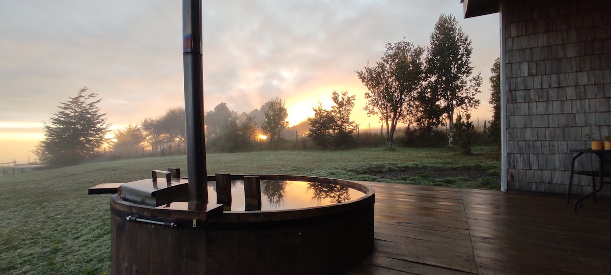 A warm wooden deck extends in front of a circular hot tub, reflecting the soft colors of the early morning sky. Lush greenery and trees frame the background, with the sun rising gently behind them, creating a serene and tranquil setting.