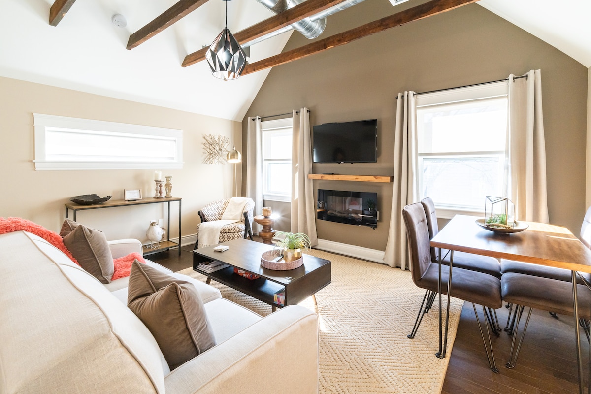 A cozy living area features vaulted beamed ceilings and large windows allowing natural light. A comfortable sofa is complemented by accent pillows, with a modern coffee table in front. An entertainment unit with a TV and a decorative fireplace create a warm ambiance near the dining area.