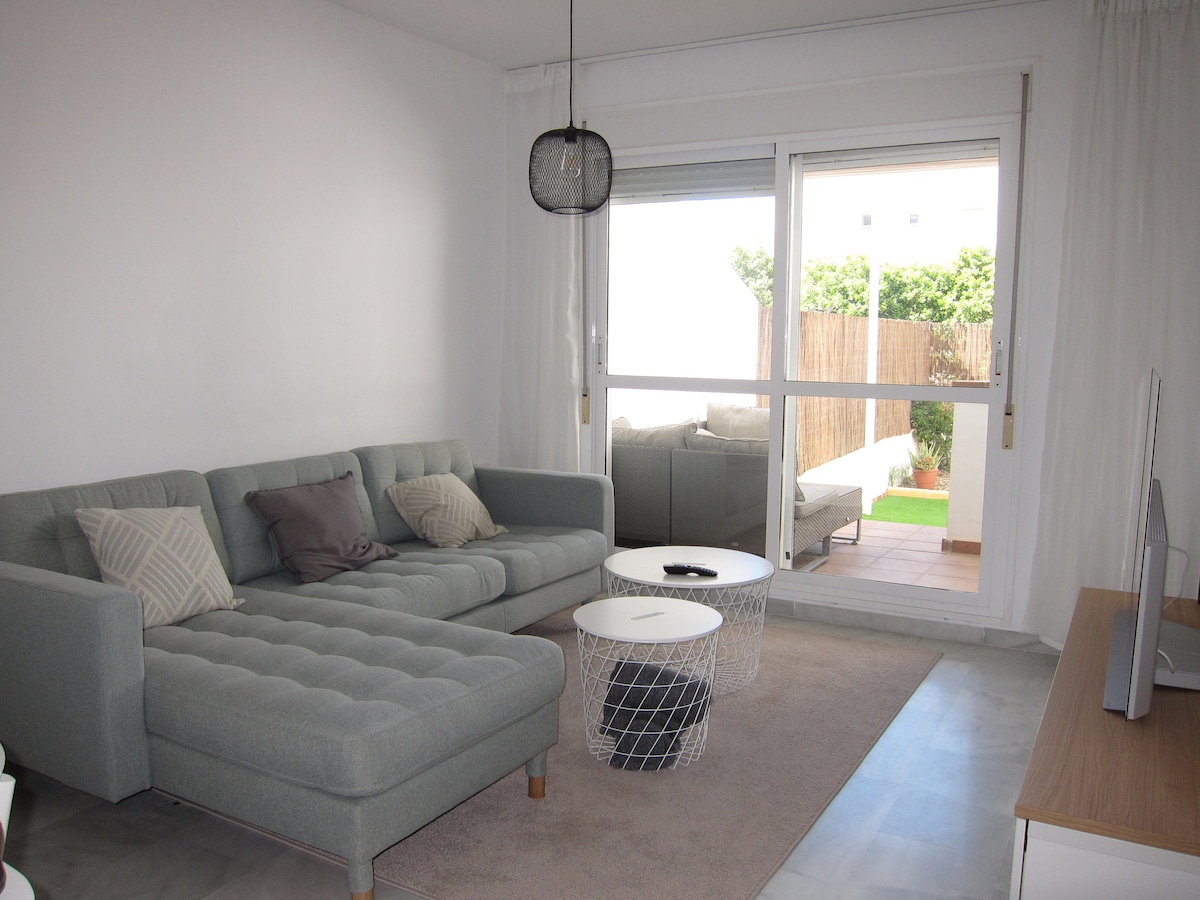 A modern living area features a gray sectional sofa with decorative cushions, placed on a soft rug. Two circular coffee tables are situated in front, while large windows allow natural light to fill the space, revealing an outdoor terrace beyond.