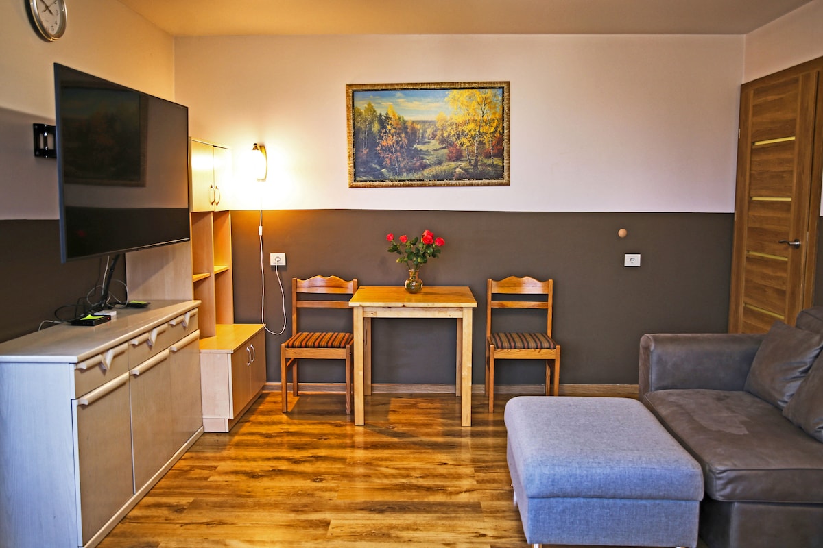 A comfortable living area features a mounted television beside a light wooden cabinet. A small dining table with two chairs sits beside a vase of fresh roses, while a plush sectional sofa offers relaxation. Soft lighting creates a welcoming ambiance.