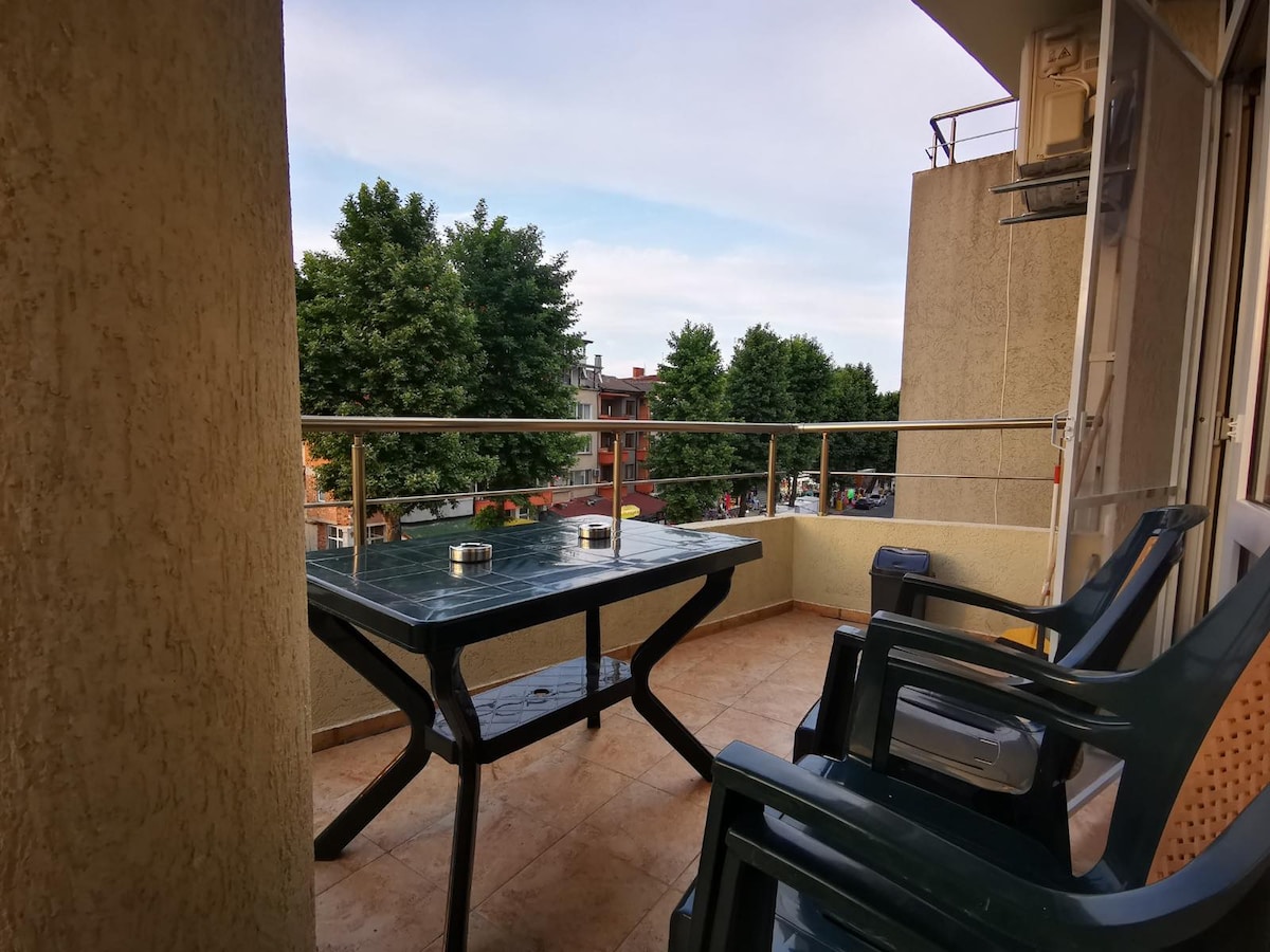 A balcony space is presented with a large table and four chairs arranged around it. Green trees are visible in the background, and the area is paved with tiles. The bright sky above complements the outdoor setting.