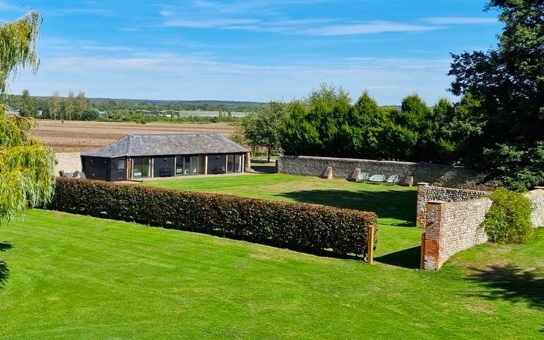 An expansive green lawn is framed by neatly trimmed hedges, leading to a modern building that features large windows. Comfortable sun loungers are positioned on the grass, inviting relaxation. The serene countryside backdrop enhances the tranquil setting, with fields visible in the distance.
