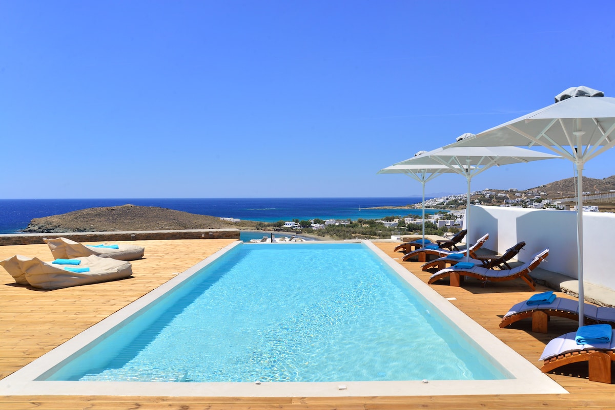 A serene outdoor area features a rectangular pool surrounded by wooden decking. Lounge chairs are arranged on one side, and large umbrellas provide shade. The expansive view includes the Aegean Sea and nearby hills, under a clear blue sky.